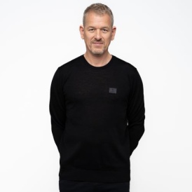 Ein Mann trägt einen schwarzen Wollpullover mit einem dunkelgrauen Patch auf der linken Brust, auf dem "1907 ELV" steht.