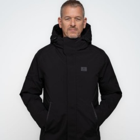 Ein Mann trägt einen schwarzen Winterparka mit hohem Kragen, durchgehendem Reißverschluss, Kapuze und Seitentaschen. Auf der linken Brust ist ein Dunkelgraues Patch auf dem "1907 ELV" steht.