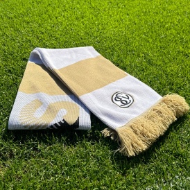 Beige-weiß gestreifter Schal mit beigen Fransen und einem SV Elversberg-Logo an dem einen Ende. In der Mitte erkennt man ein großes beiges "S". Der Schal liegt gefaltet auf einem Rasenfeld, sodass man nicht das gesamte Design erkennen kann.